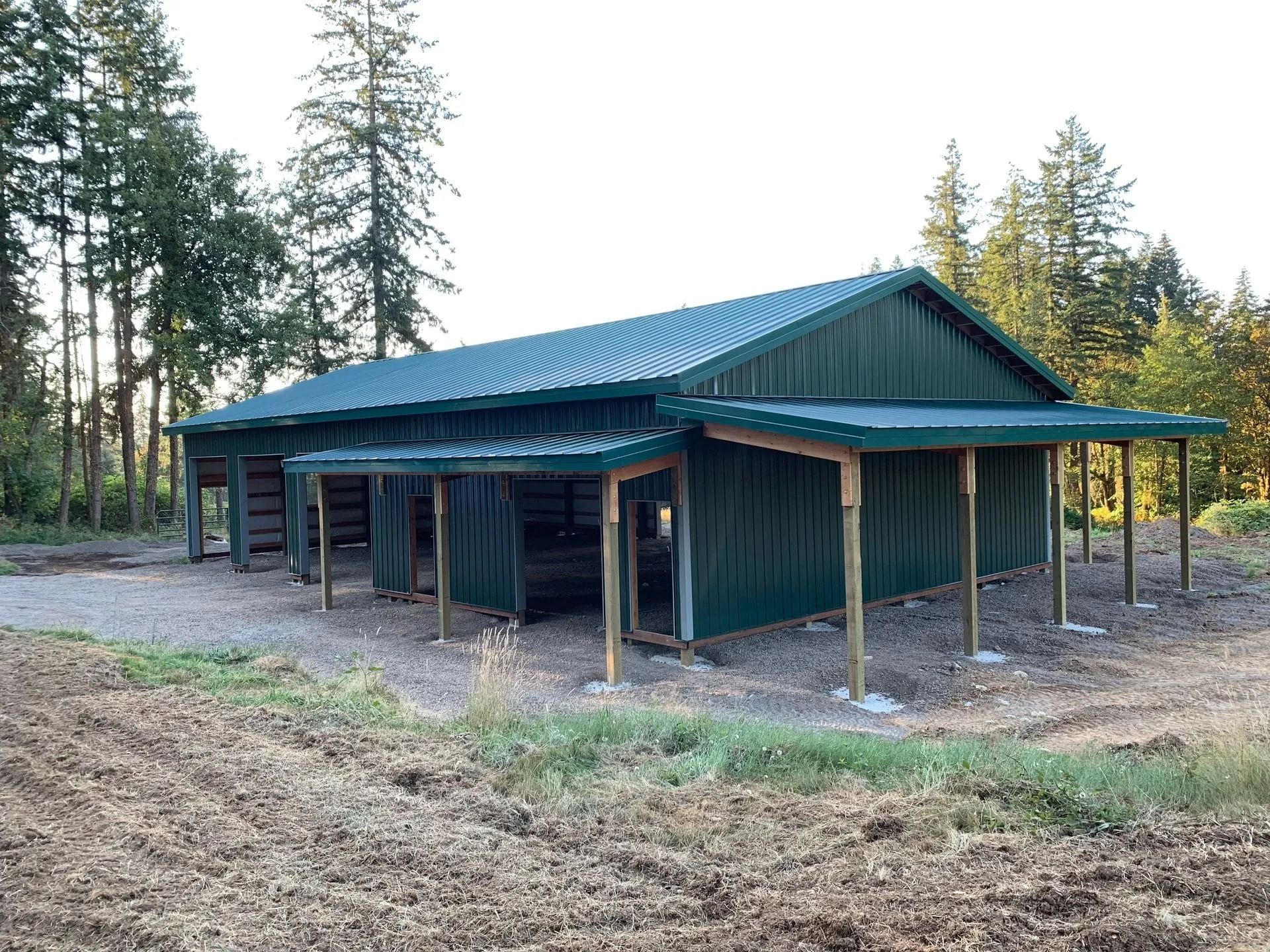 Green metal barn in a field with wooden support posts and a porch-like overhang.