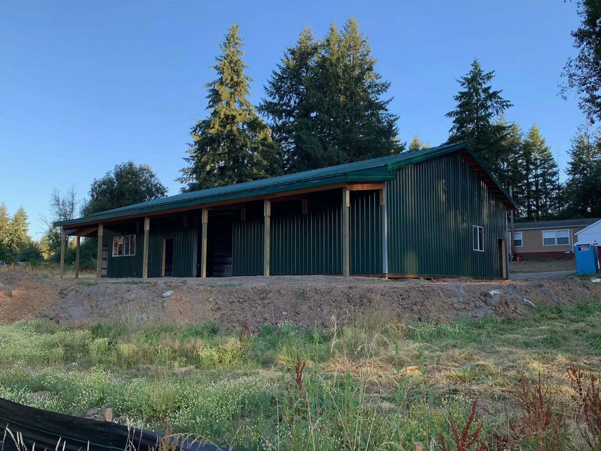 Green metal-clad building with a covered porch and gravel base, in a wooded area under a blue sky.