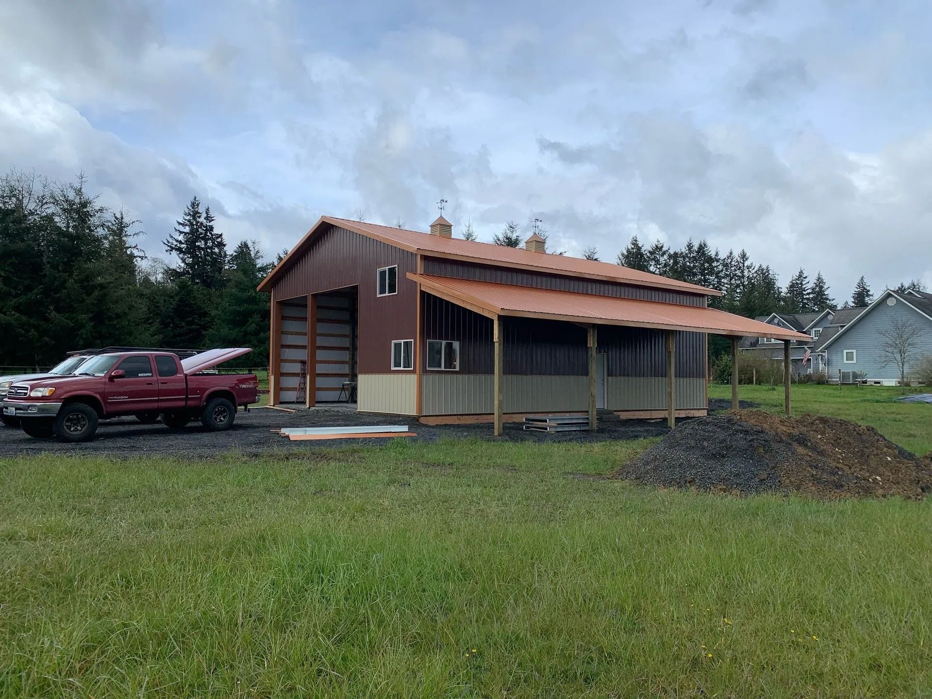 Red pickup truck parked near a large brown barn with a screened porch and brown metal roof, on a grassy lot.
