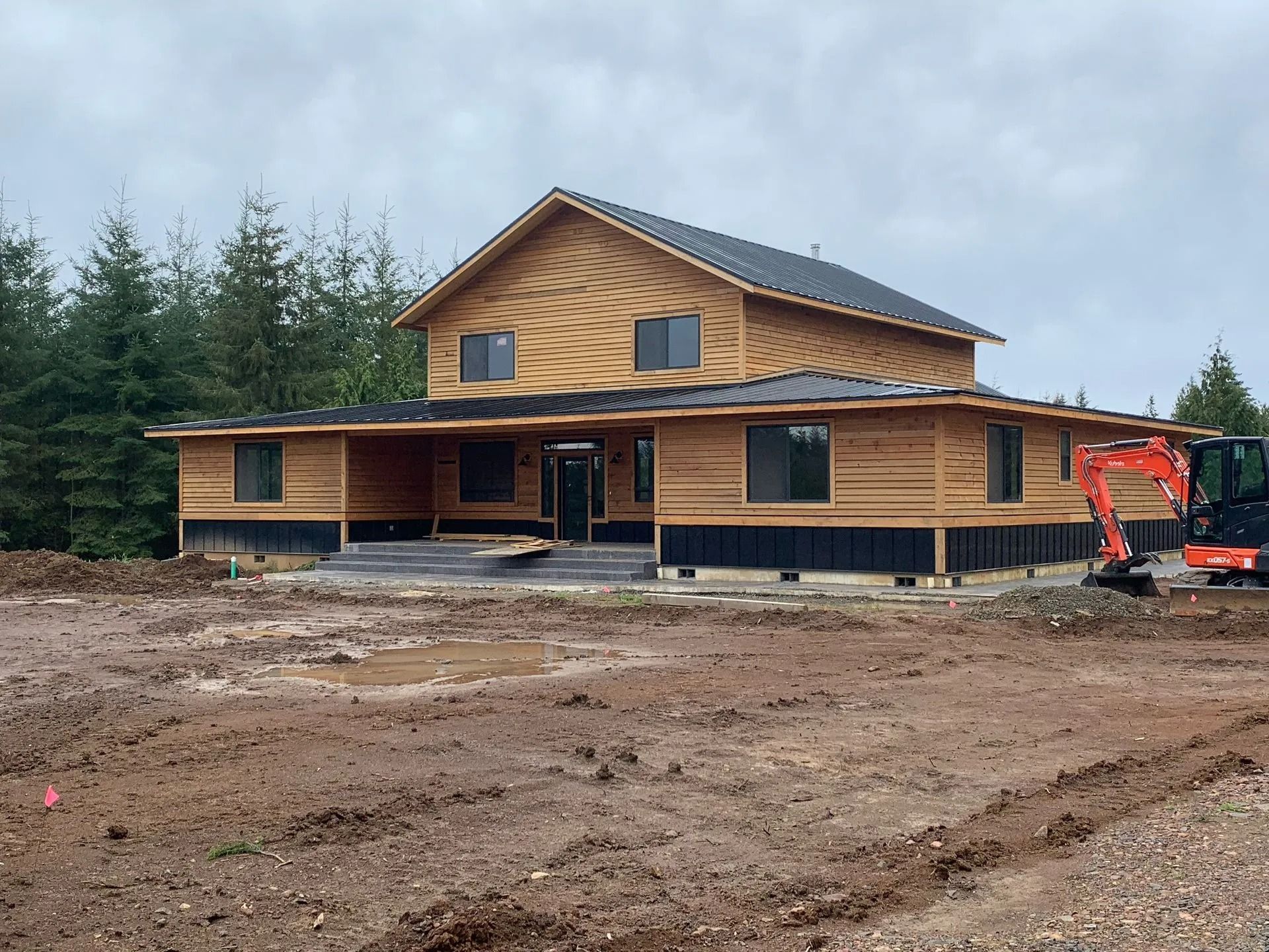 House under construction; wood siding, two stories, front porch, and construction equipment on muddy ground.