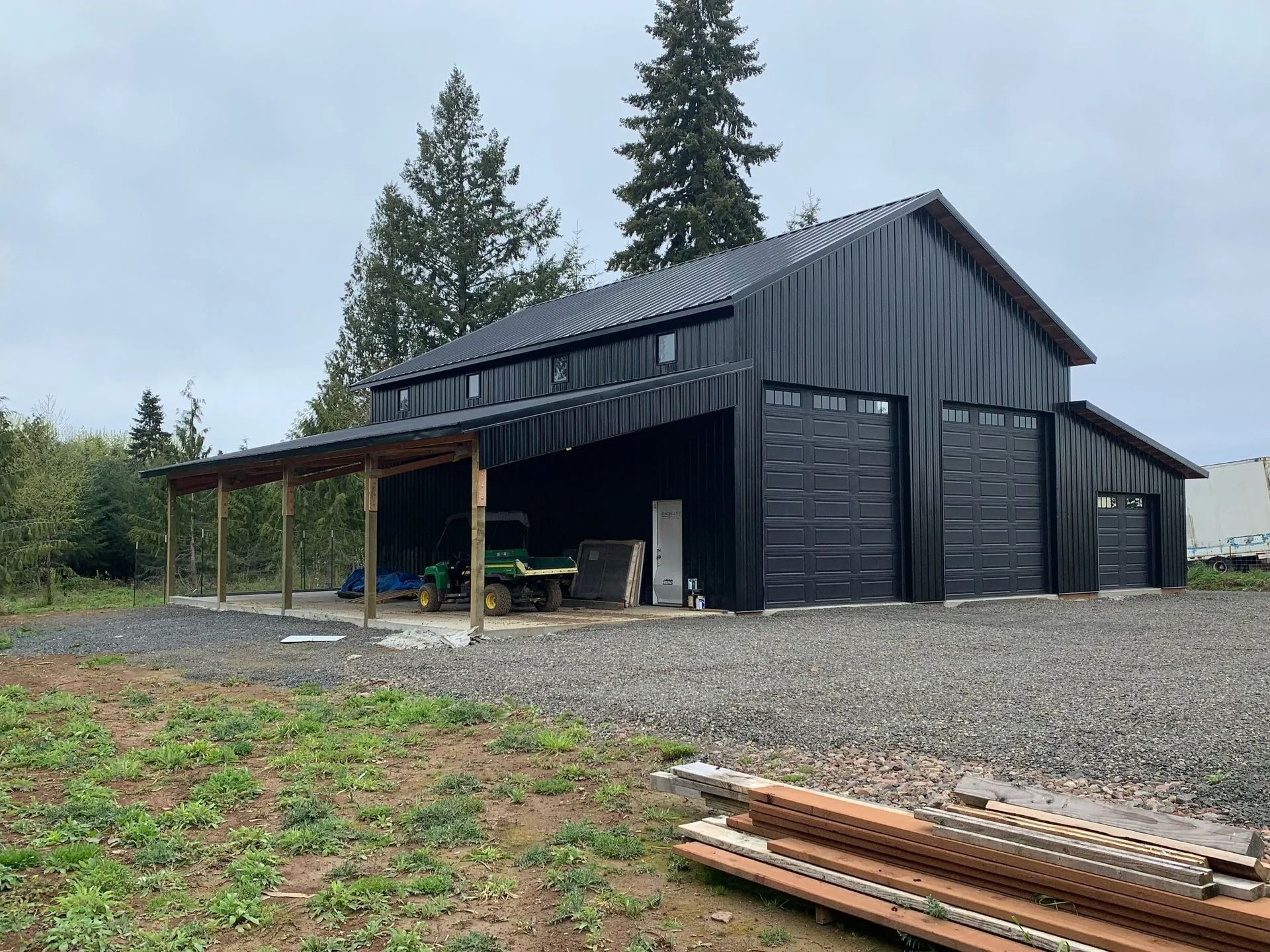 Black barn with two garage doors and a carport. Gravel driveway and green grass in front. Overcast sky.
