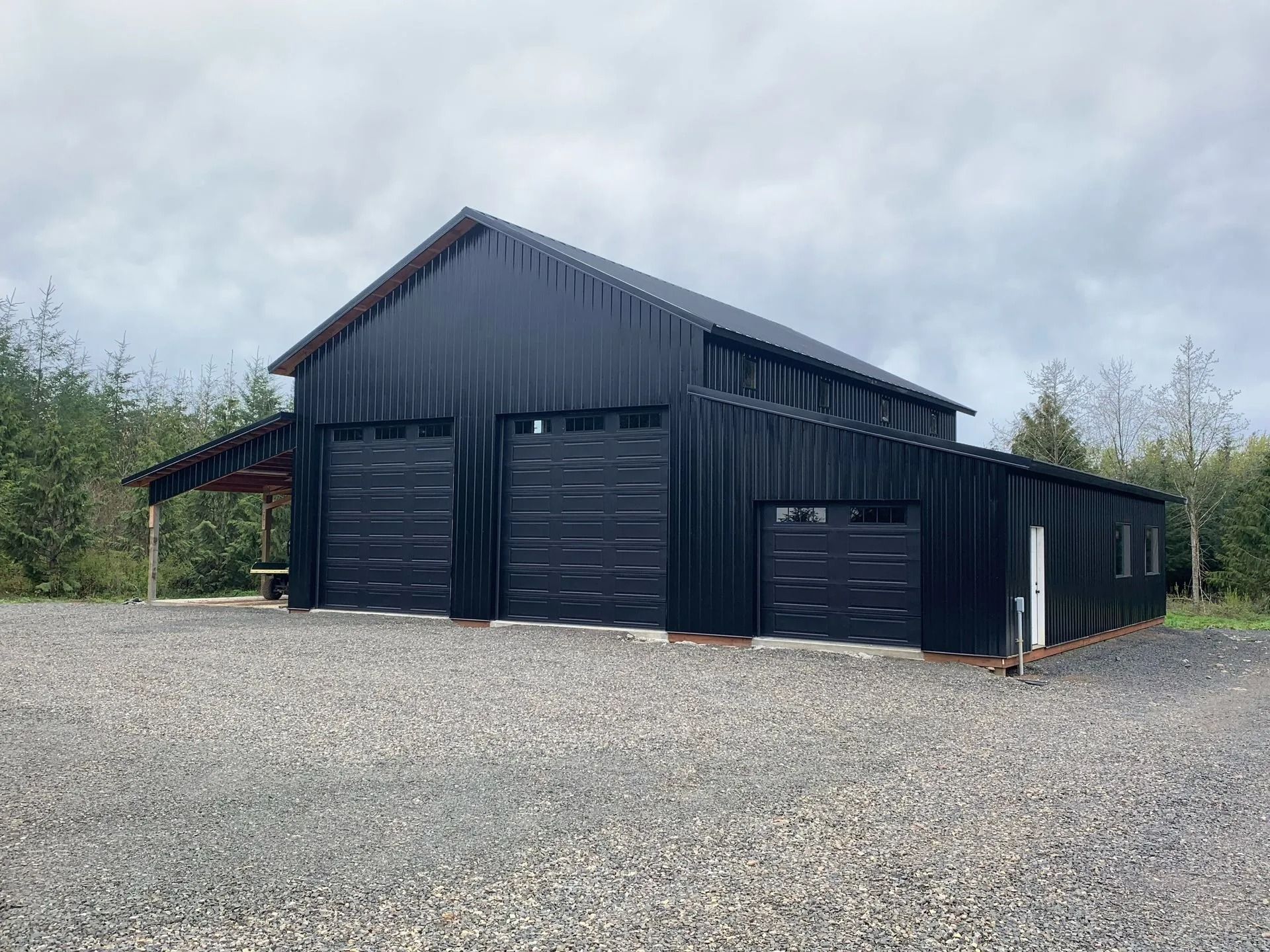 Black barn-style building with three garage doors and a gravel driveway under a cloudy sky.