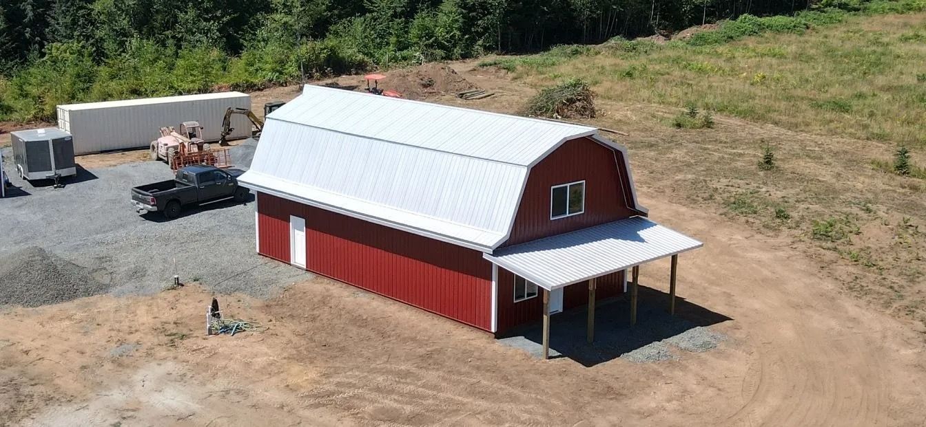 Red barn with a white roof and porch, set in a gravel lot with a pickup truck and trees.