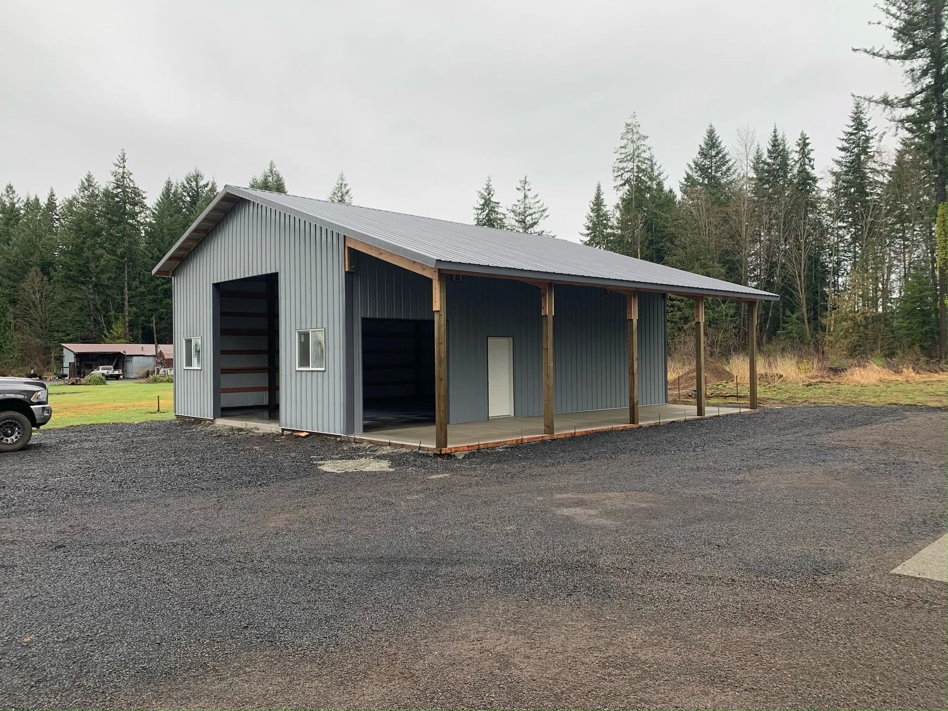 Gray metal barn with a covered porch; gravel driveway, trees in background.