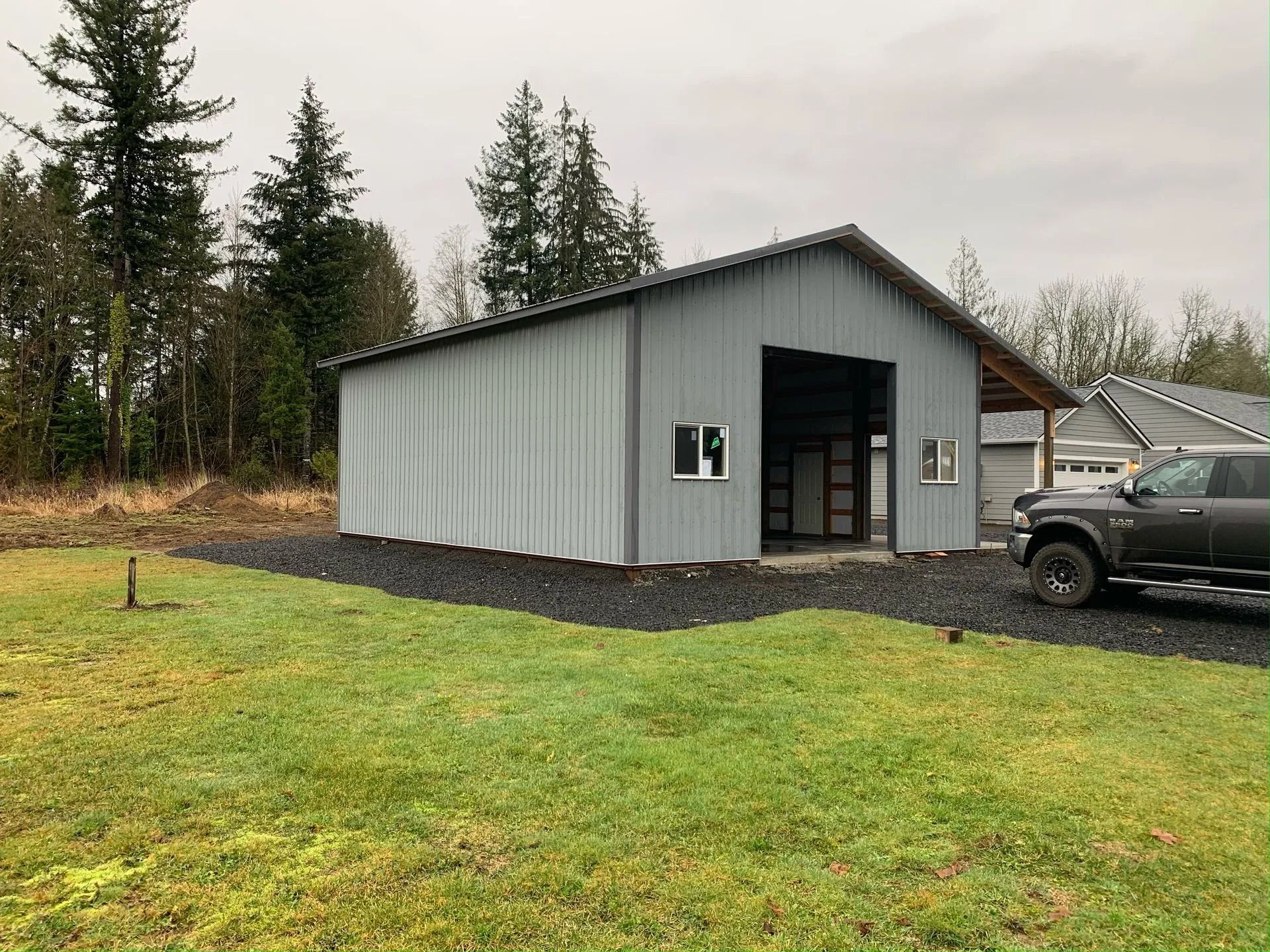 Gray metal workshop with large open door, two windows, parked truck, surrounded by gravel and grass.
