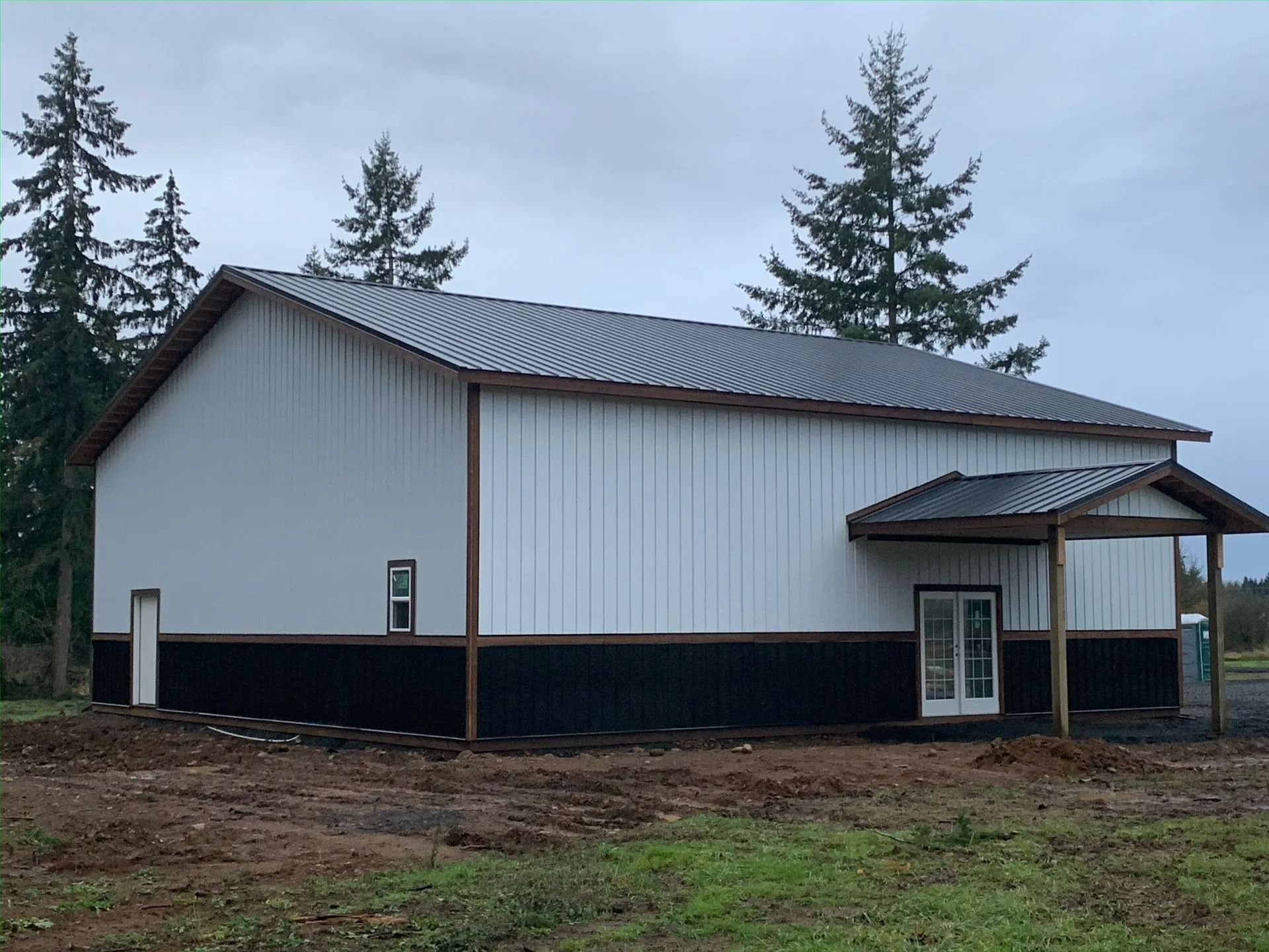 White and black metal building with a covered entrance, surrounded by trees and a cloudy sky.