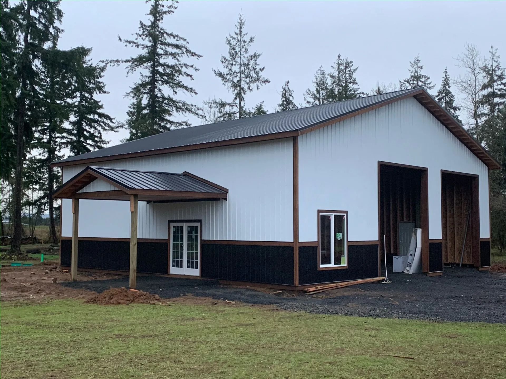 White and black metal building with a covered porch and two garage doors, set on gravel.