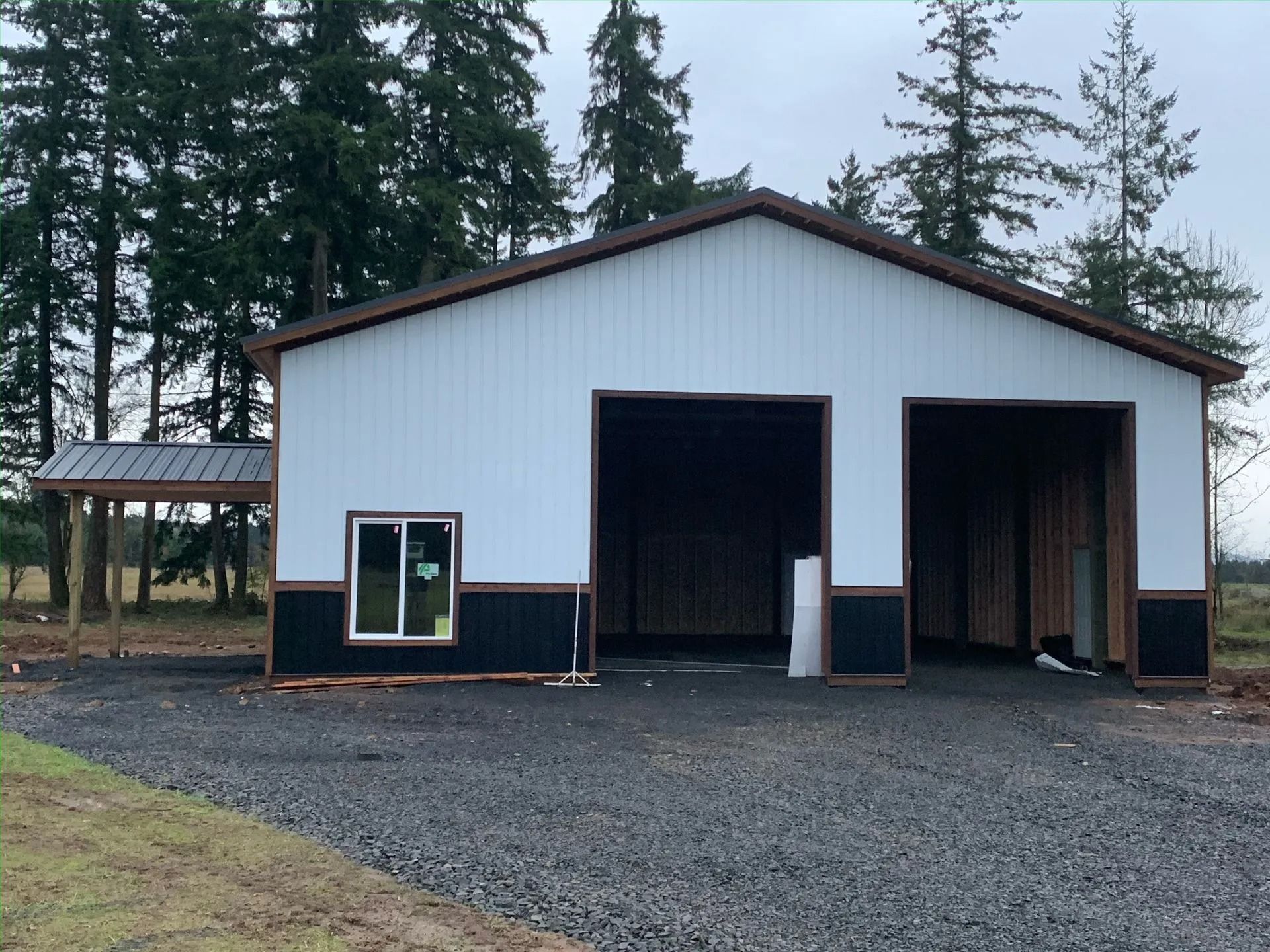 White and black metal garage with two bays, small window, brown trim, gravel driveway, and trees.