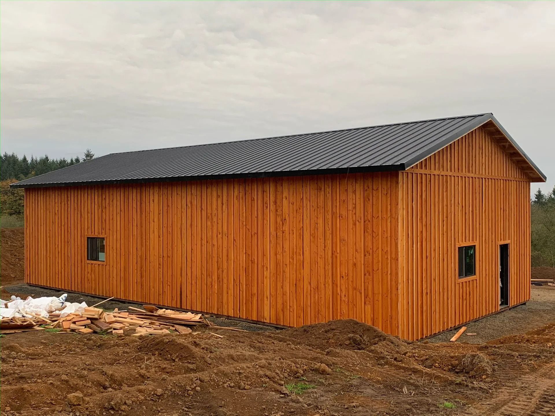 Wooden barn with dark roof, two small windows, and a doorway. Set on a dirt lot under an overcast sky.