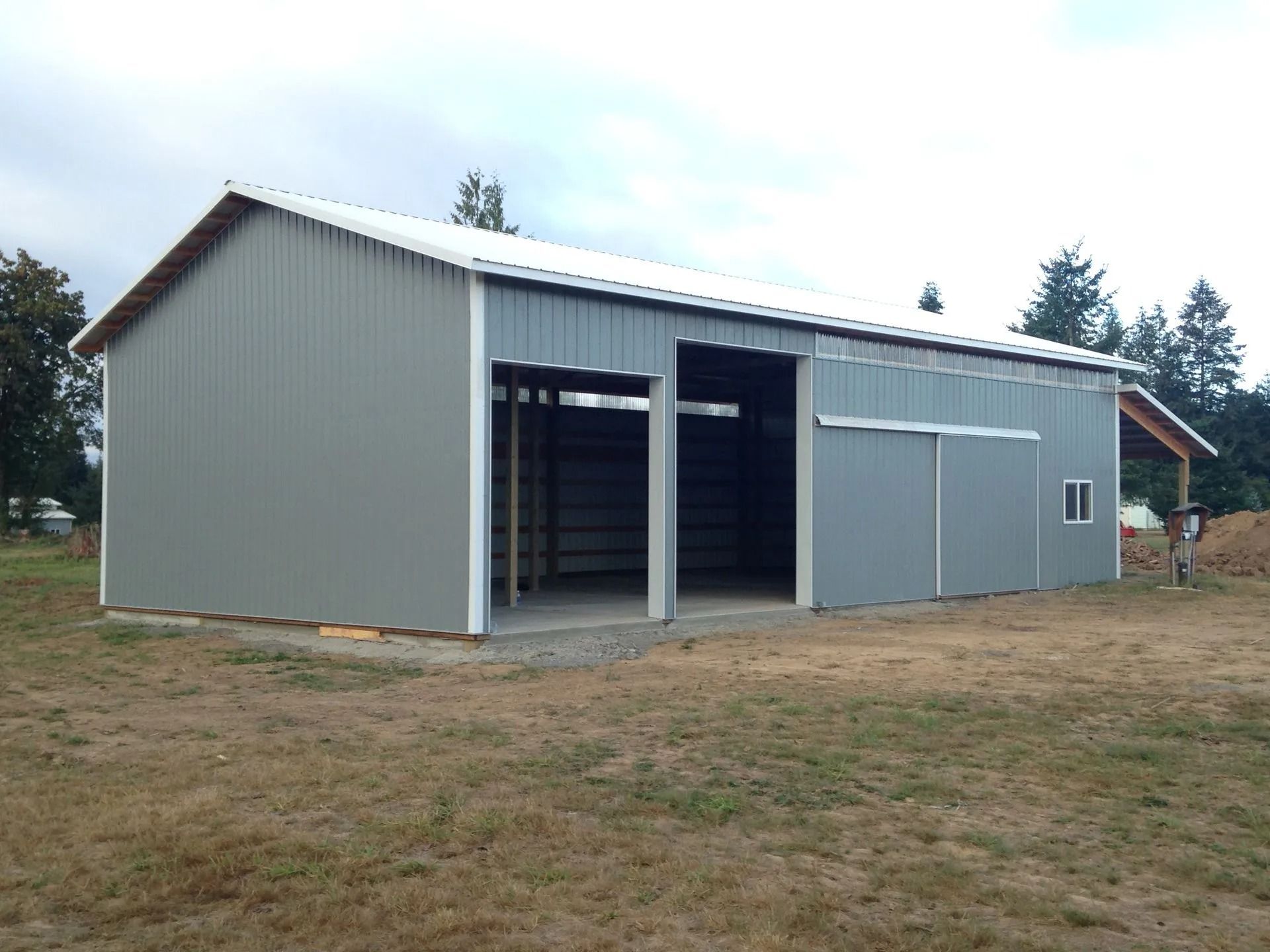 Gray metal barn with open bays, sliding door, and a white roof on a grassy lot.