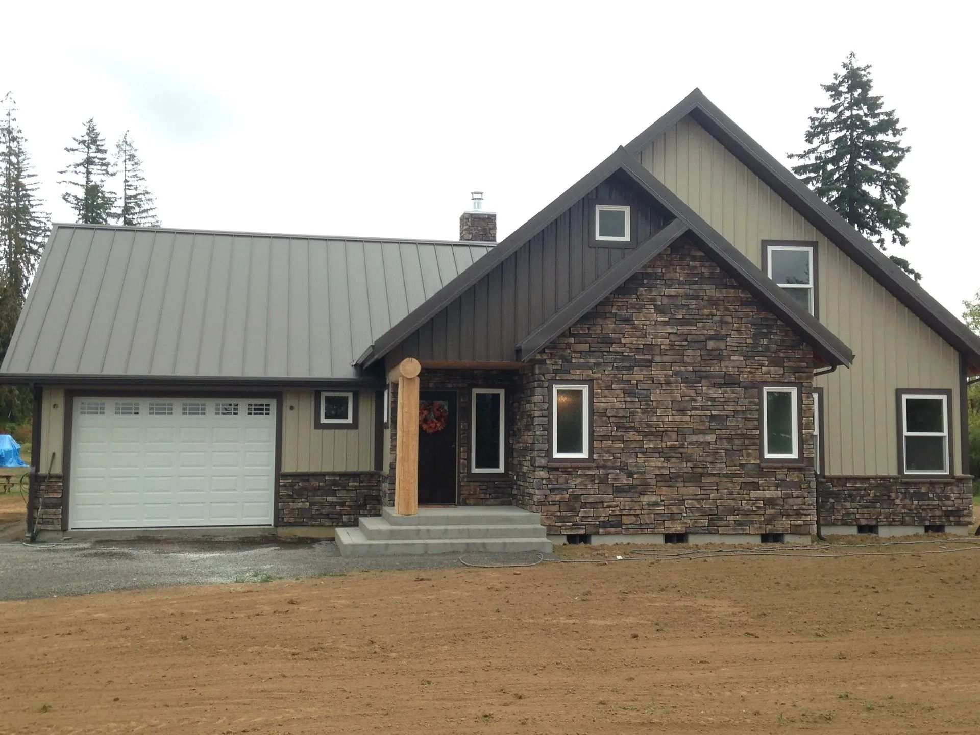A two-story house with a stone facade, beige siding, and a metal roof. A garage door is visible.