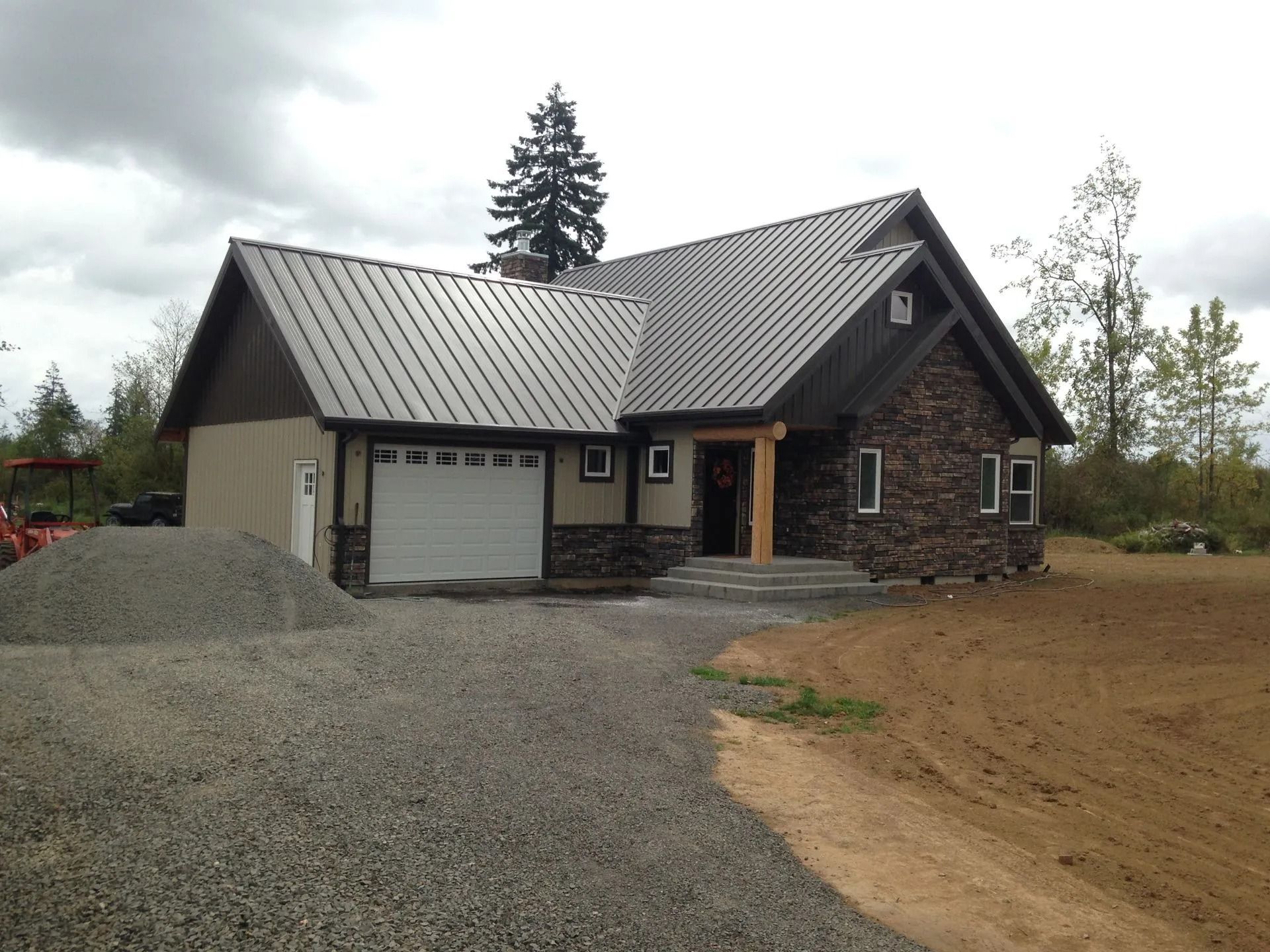 New house with metal roof, stone accents, and gravel driveway under a cloudy sky.