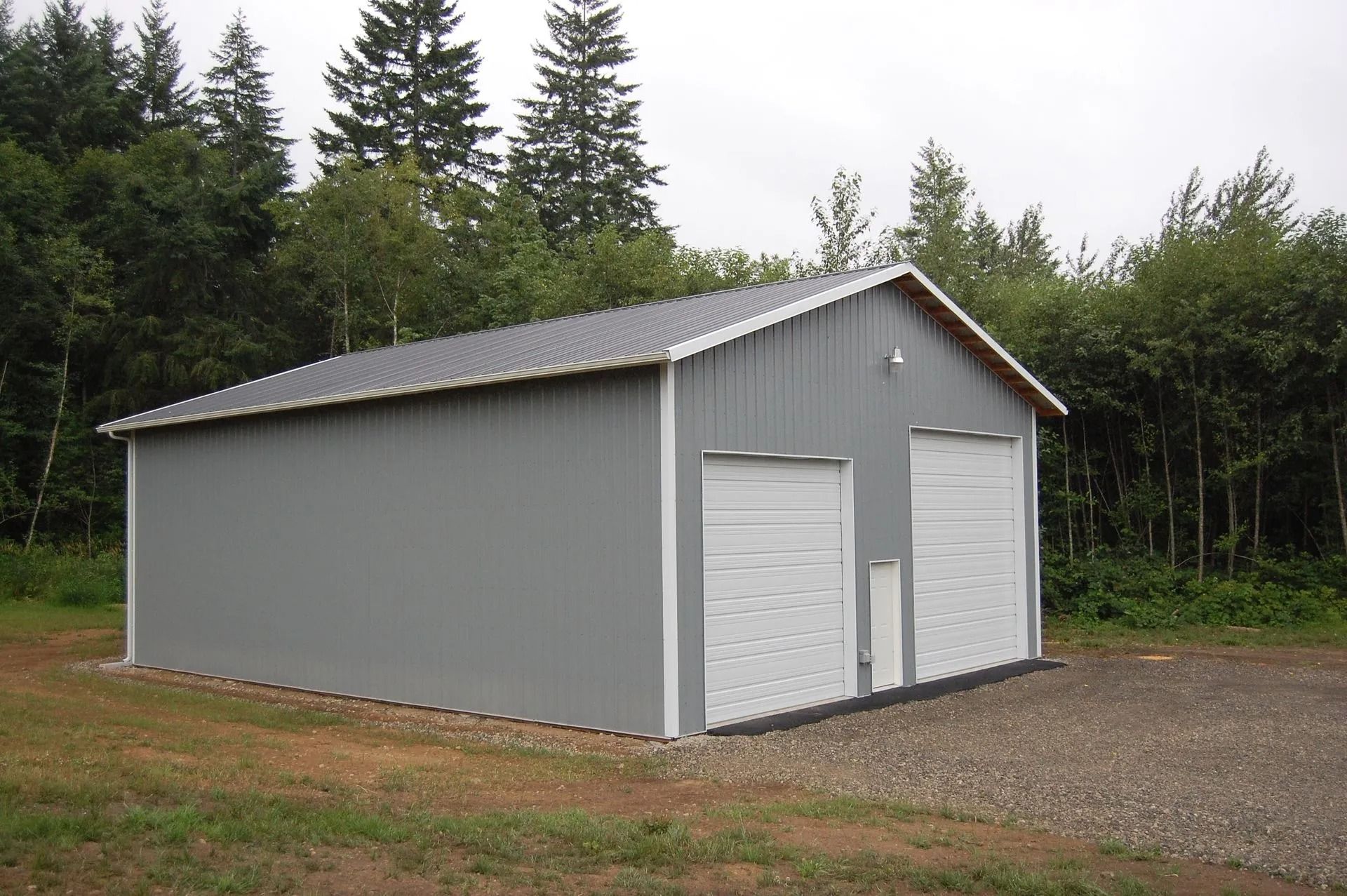 Gray metal garage with two white doors, set on gravel, trees in the background.