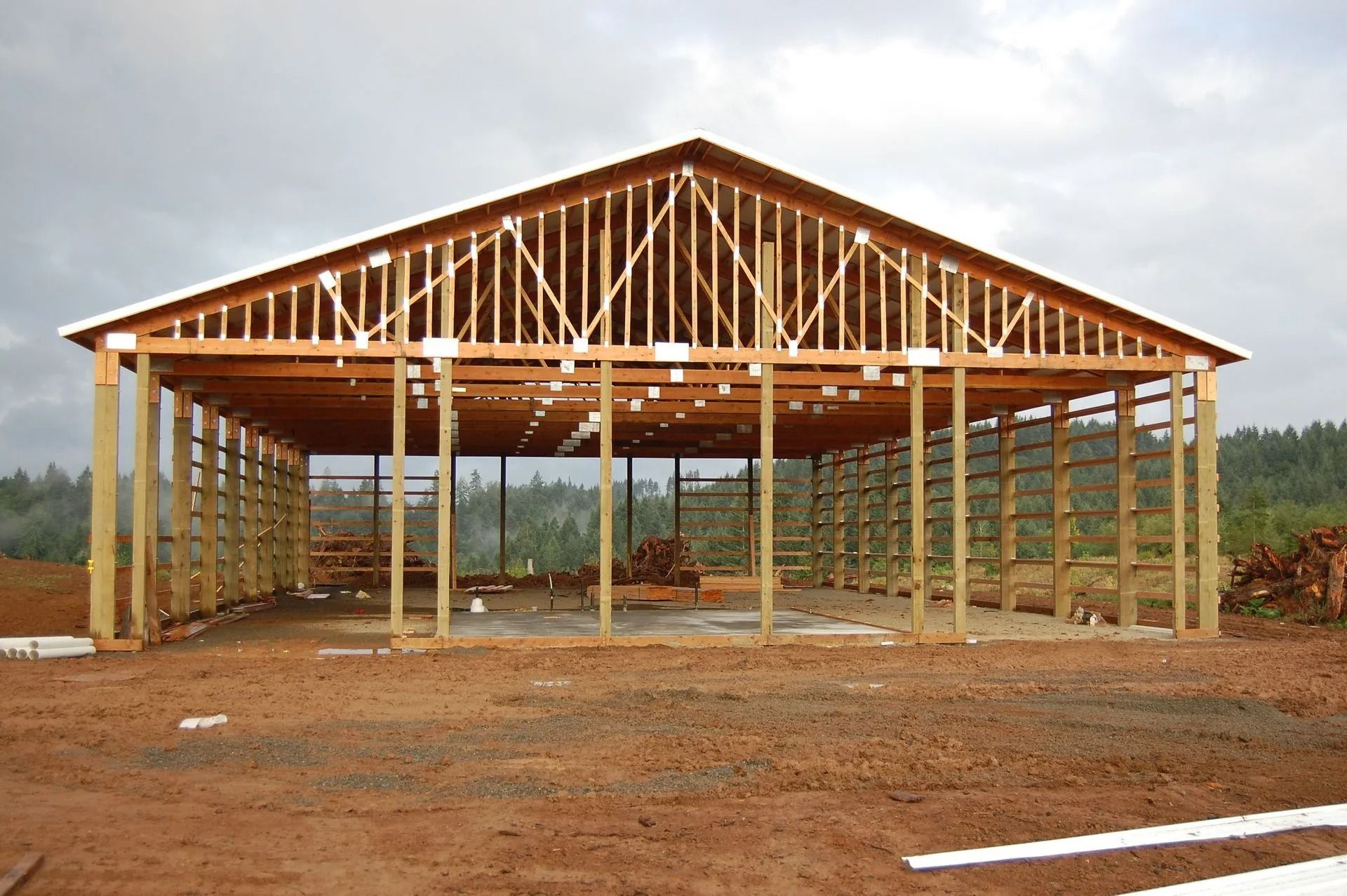 Wooden barn frame under construction, set on dirt with cloudy sky in the background.