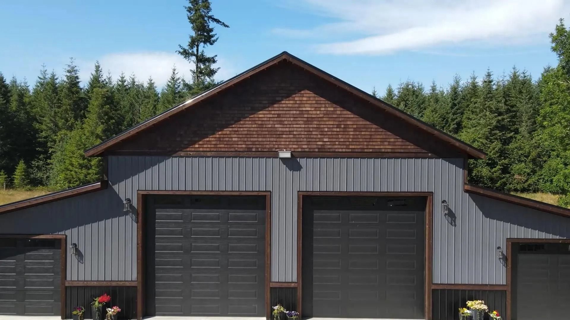 Gray garage with black doors and brown trim under a wooden gable roof, set against trees.