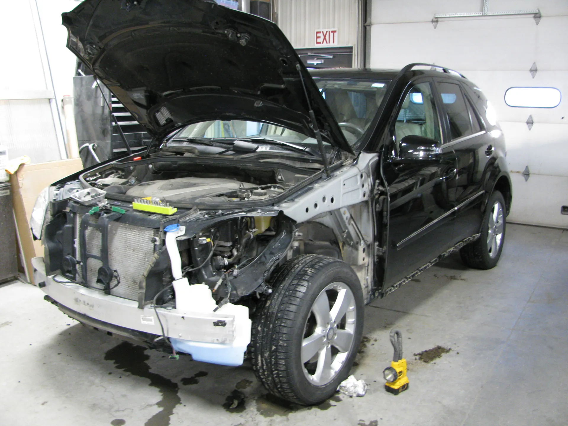 Black SUV in a repair shop with front end damage, hood open, parts removed.