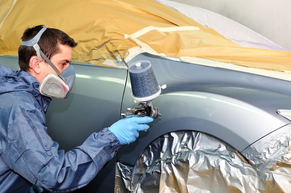 Person in protective gear painting a car's fender with a spray gun in an auto body shop.