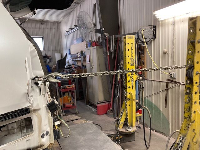 A damaged white vehicle being repaired with a chain and yellow pulling frame inside a garage.