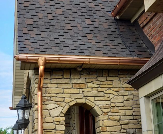Stone-front house with dark shingled roof, copper gutters, and arched doorway under blue sky