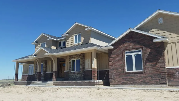 Large tan and brick house with a covered porch and multiple dormer windows on a sunny, unfinished lot.