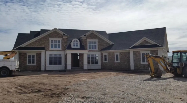 Large stone house under construction with a front porch, windows, and construction equipment in the yard