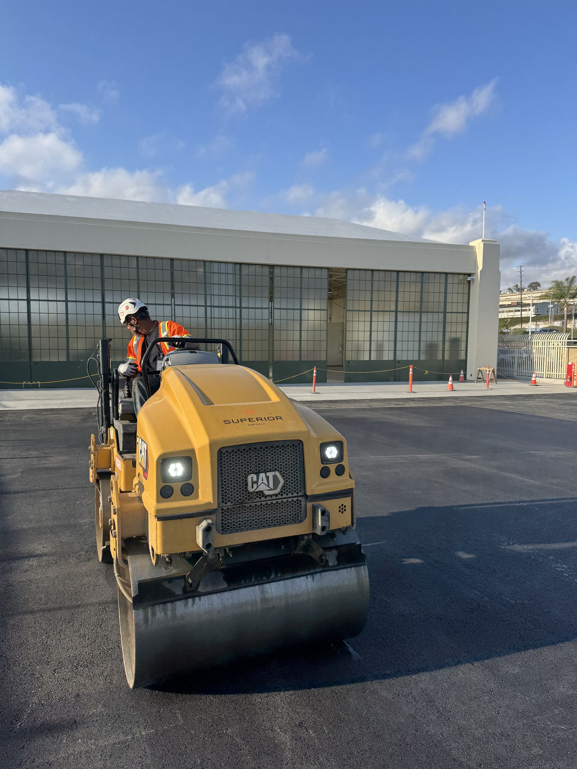 Yellow road roller on a paved lot with a worker in orange safety gear in front of a building