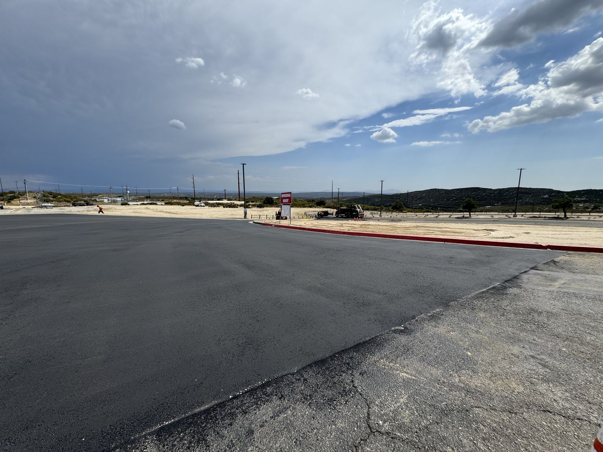 Empty paved lot beside a road under a cloudy sky with hills in the distance