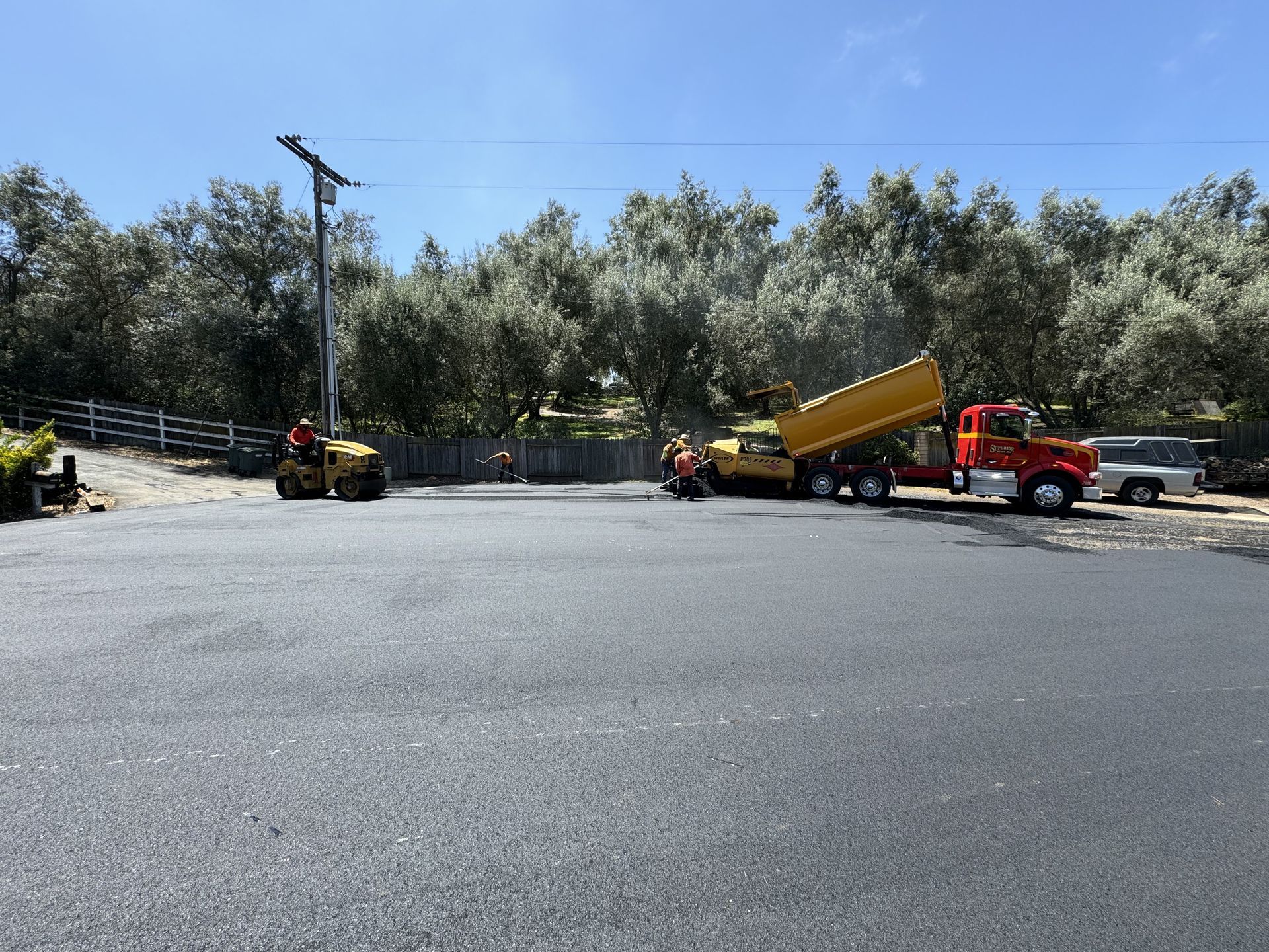 Construction vehicles on a paved lot, including a red truck unloading yellow material under a blue sky.