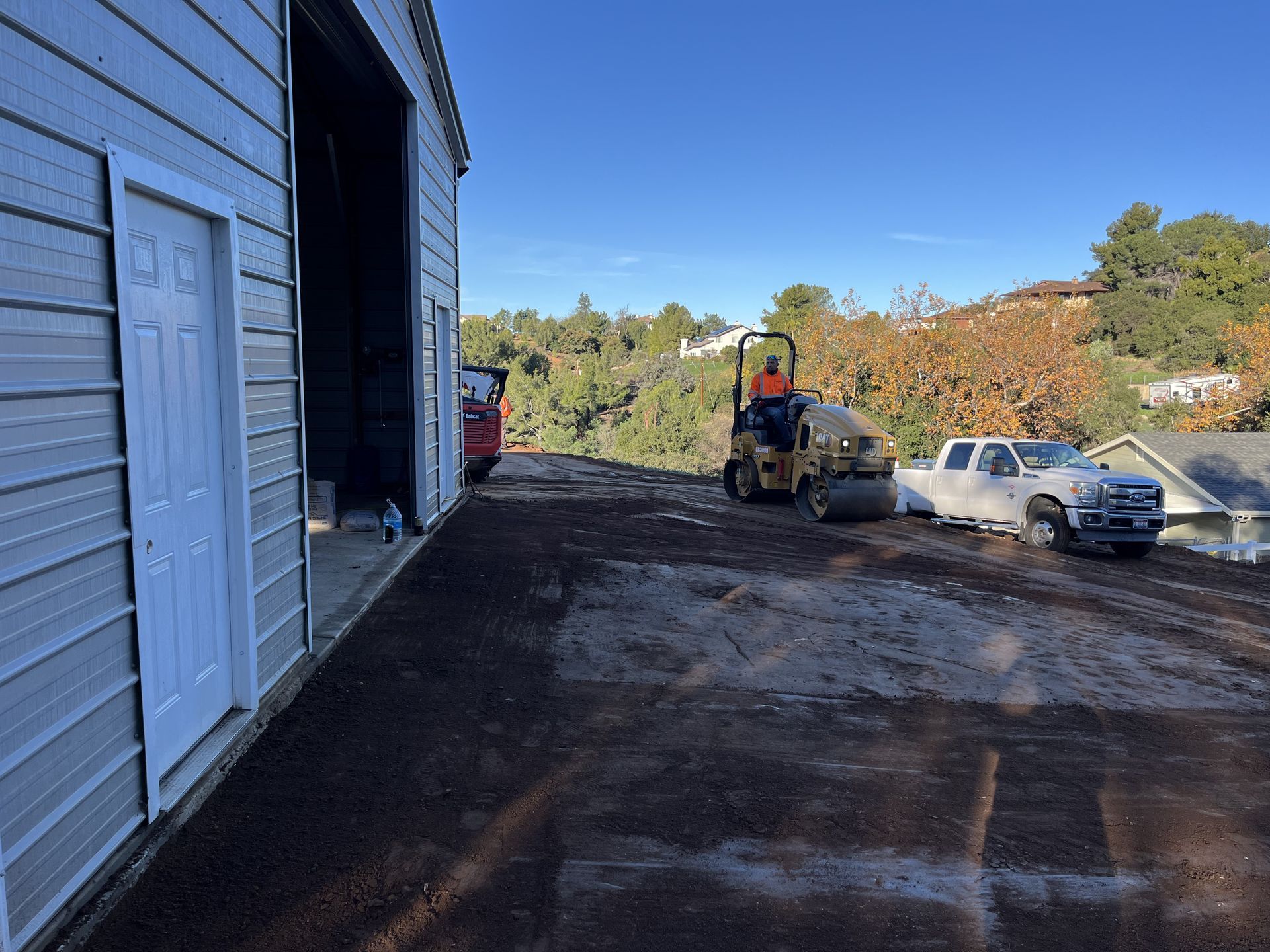Construction site beside a blue building, with a small loader and white pickup on a dirt lot.