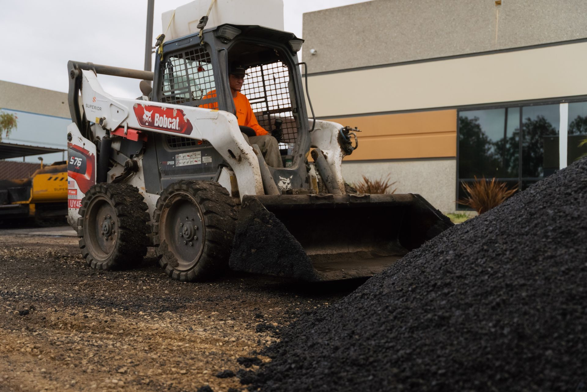 White skid steer loader scoops black gravel beside a building at a worksite