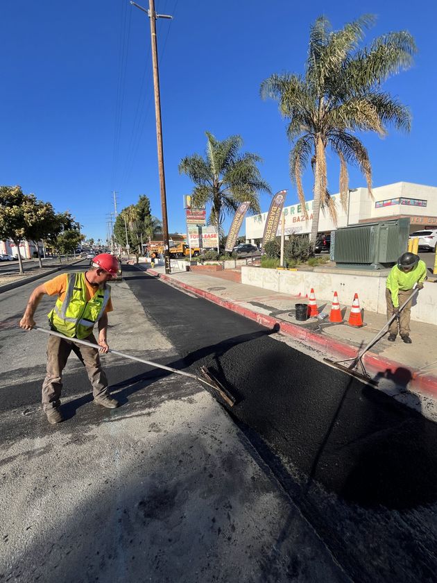 Roadwork crew paving a street, with workers beside fresh black asphalt and traffic cones under a blue sky