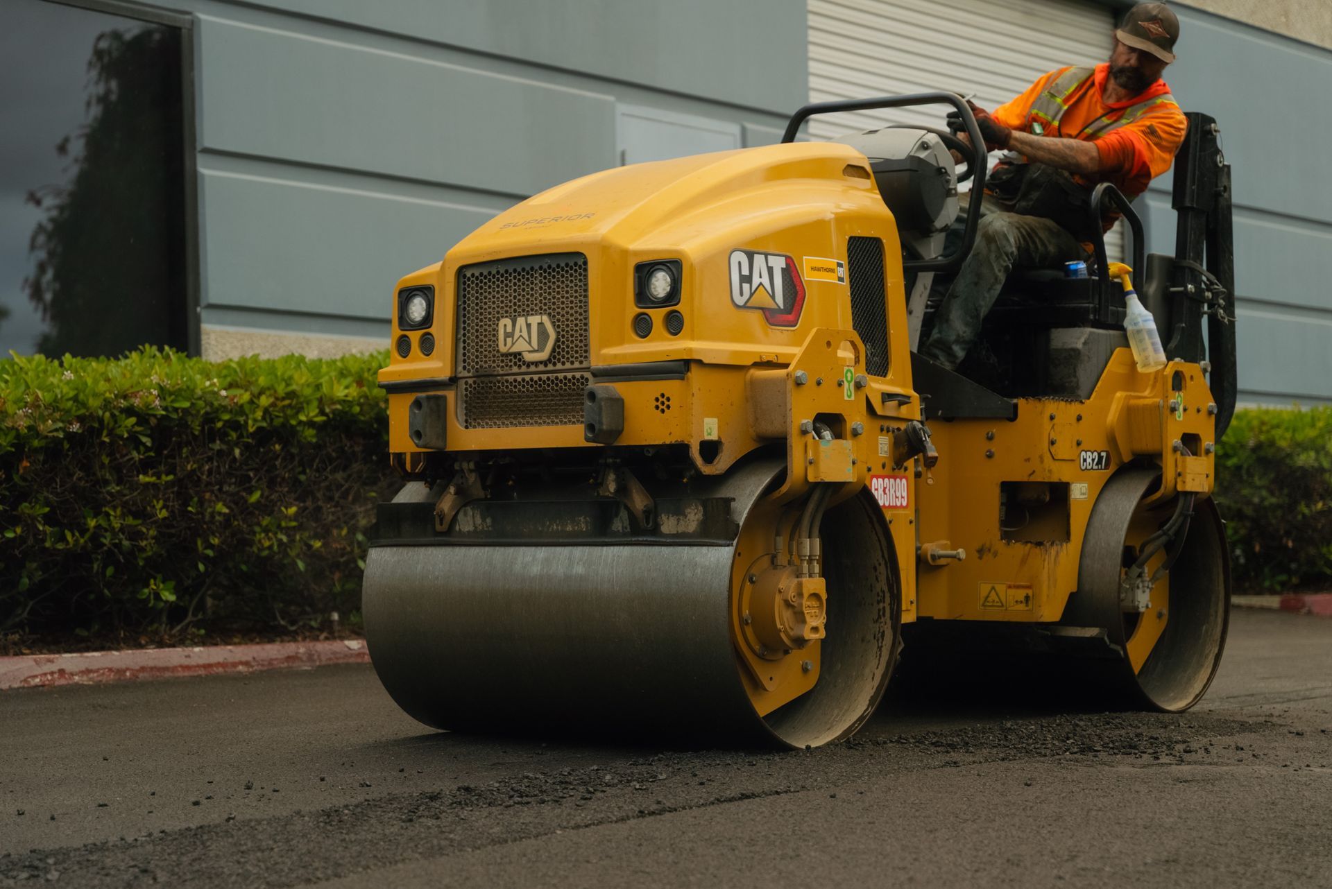 Yellow road roller with a worker in orange vest compacting pavement outdoors
