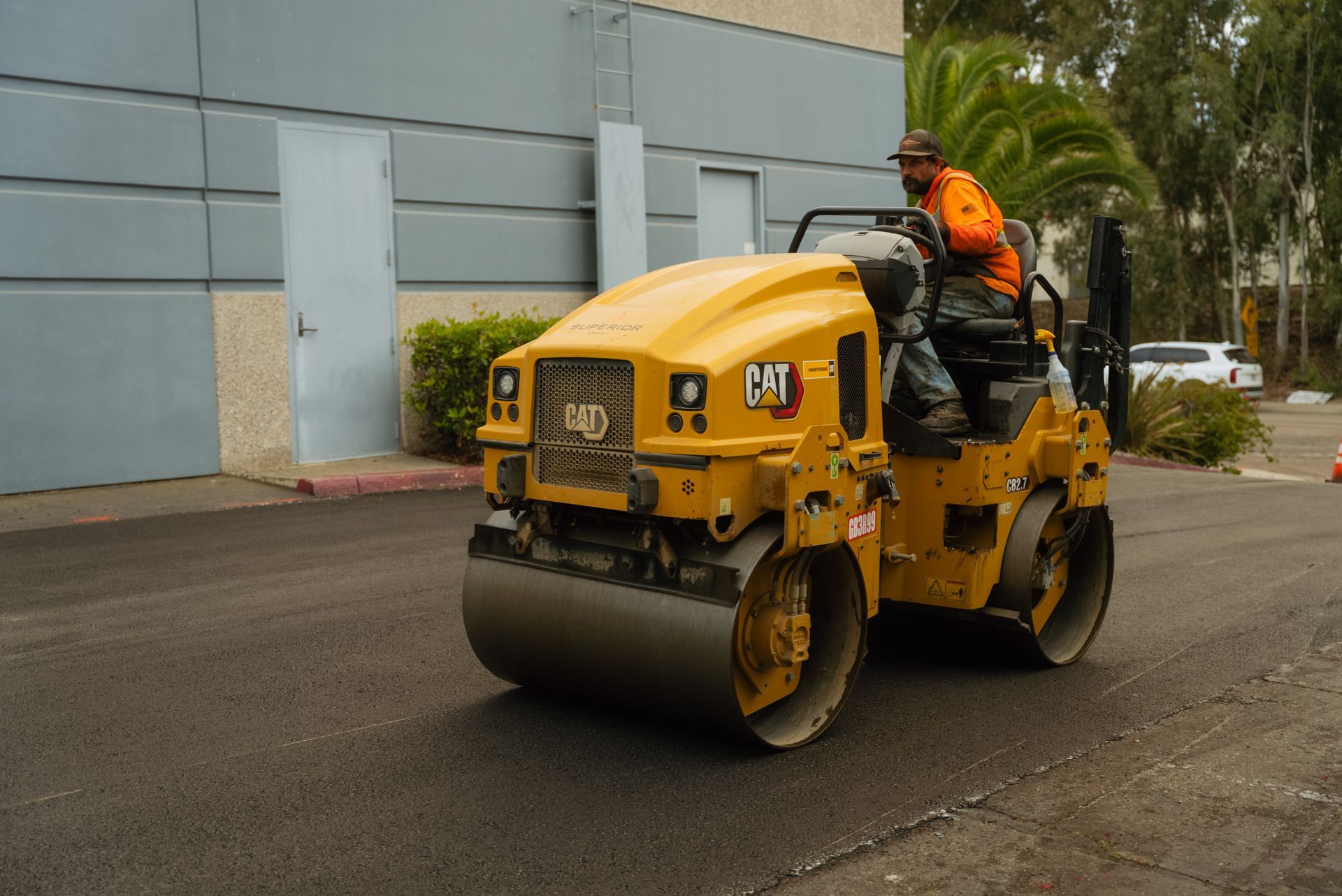 Worker on a yellow road roller compacting pavement beside a building.