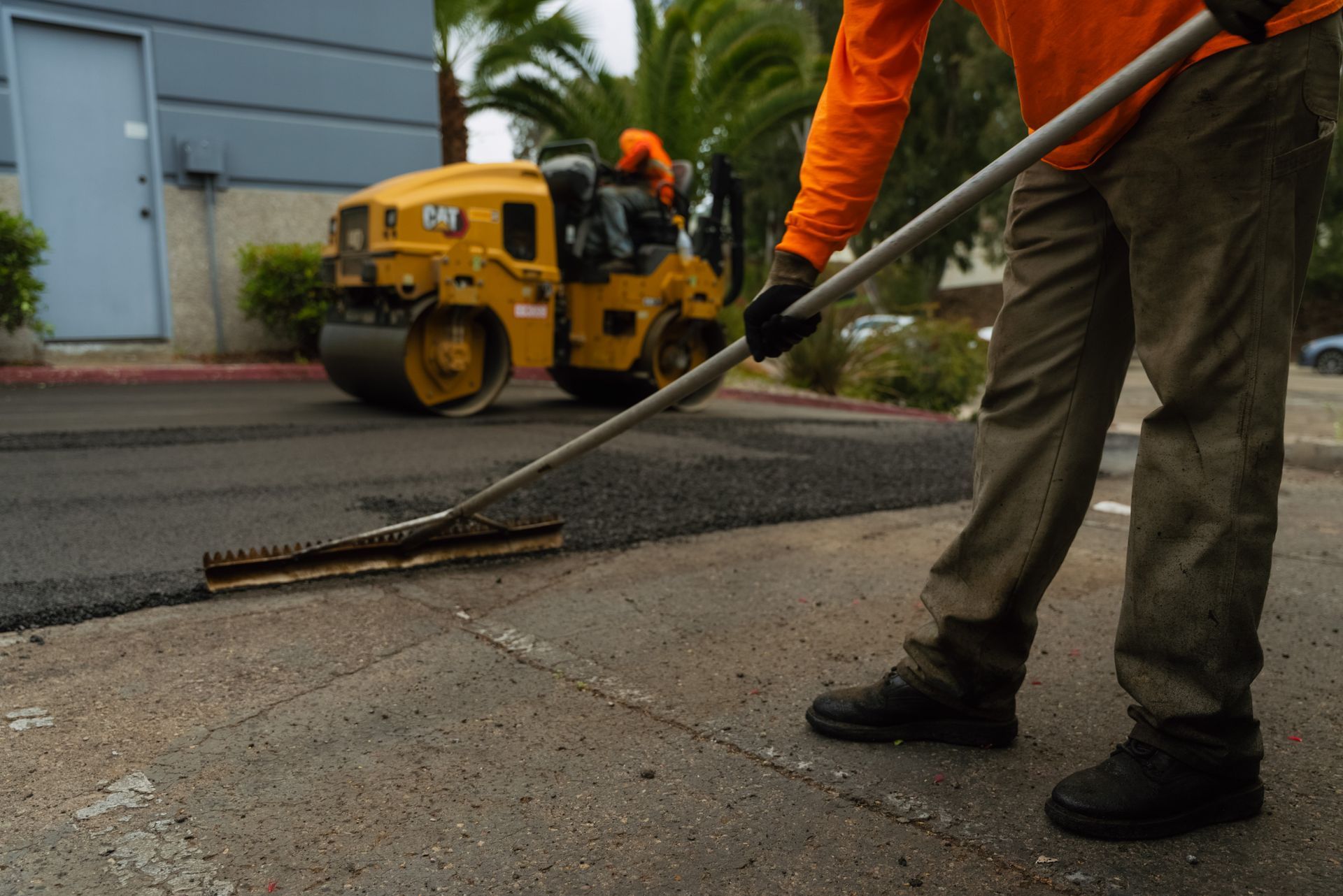 Road worker power-washing pavement beside a yellow road roller on a wet street.