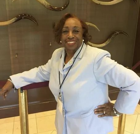 Smiling woman in a light blue suit stands near a gold railing. Dark background with decorative horns.