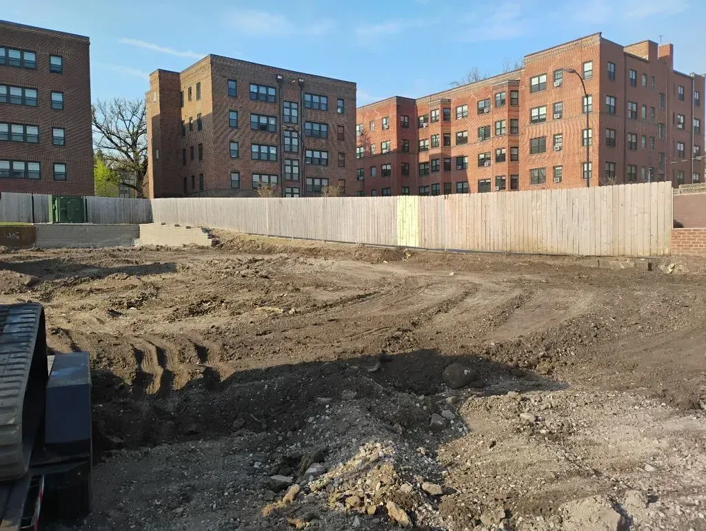 Construction site with dirt, wooden fence, and brick apartment buildings.