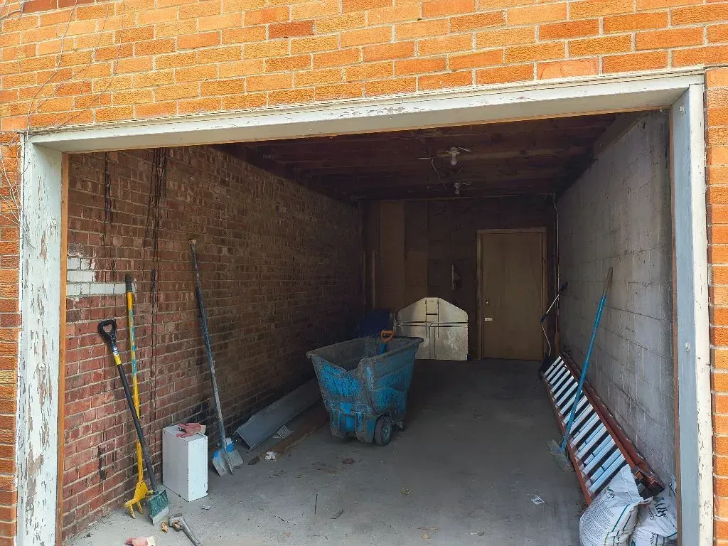 Open doorway into a brick and concrete storage area with tools, a wheelbarrow, and wooden door.