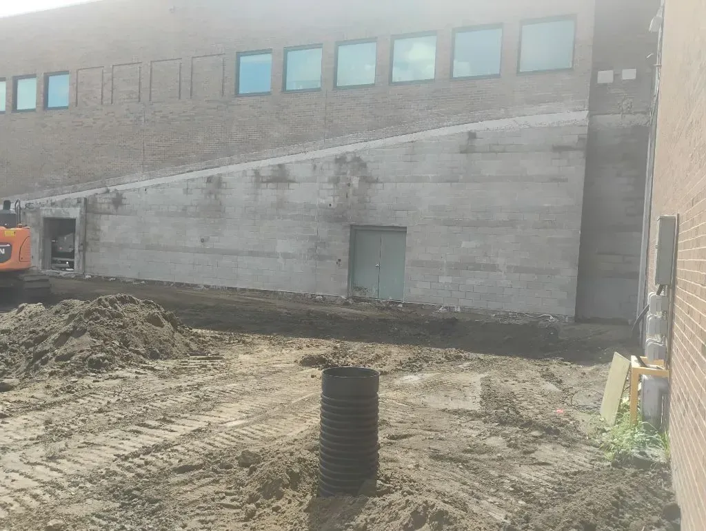 A building exterior with a trench, dirt, and a cylindrical black pipe. An excavator is partially visible.