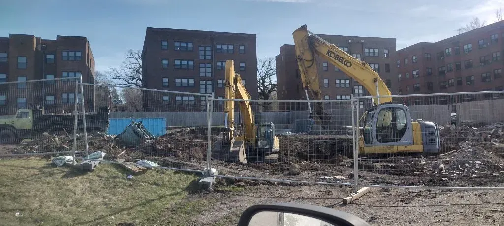 Construction site with two yellow excavators behind a fence, working on dirt. Buildings in the background.