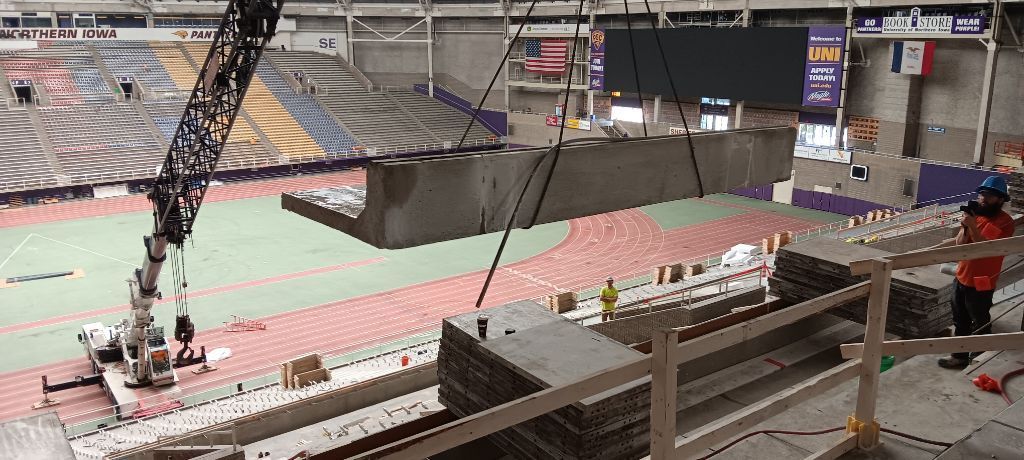 A large crane lifting a concrete barrier inside a stadium; construction in progress.
