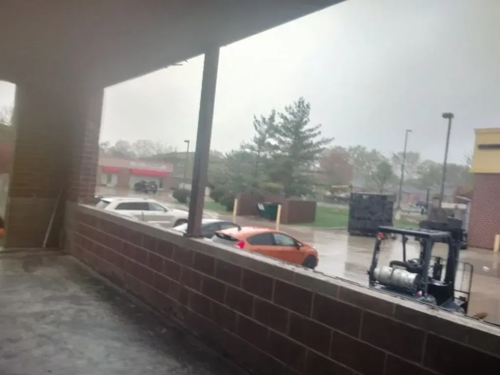 View from under a covered porch, rain falling on a parking lot with cars and a small construction vehicle.