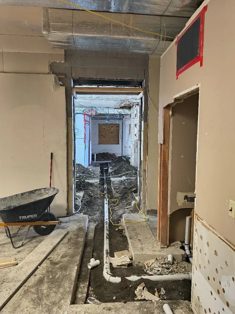 Hallway under construction, dirt and debris line the floor, wheelbarrow on the left, exposed walls, doorway to another area.