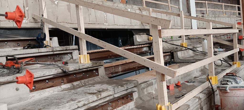 Wooden safety railing on a construction site, with orange and yellow supports, and exposed concrete.