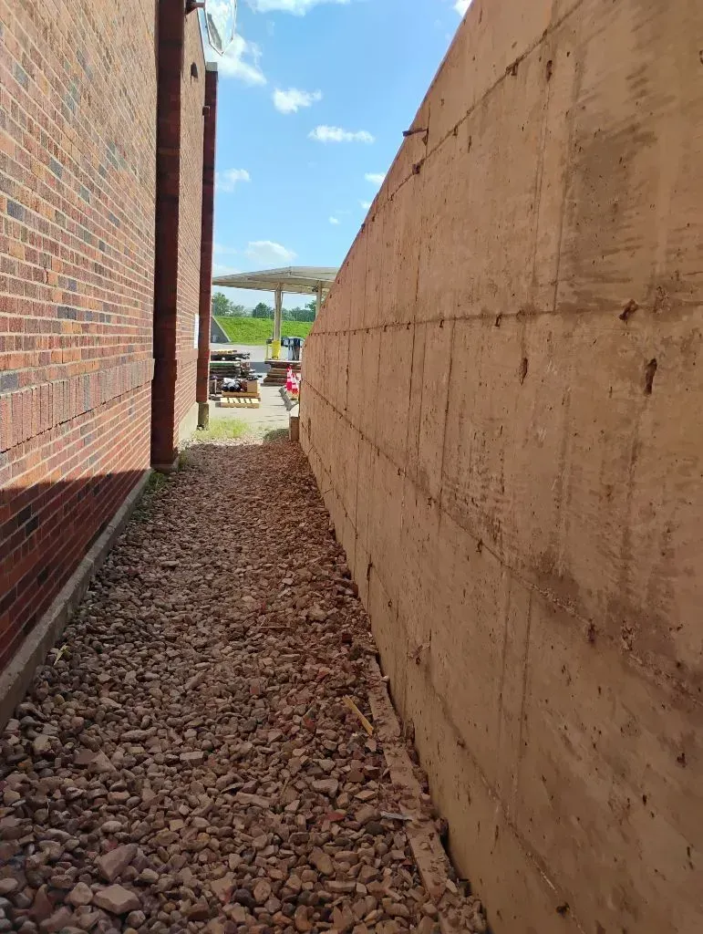 Narrow gravel path between a brick building and a light-colored concrete wall; sunny day with blue sky.