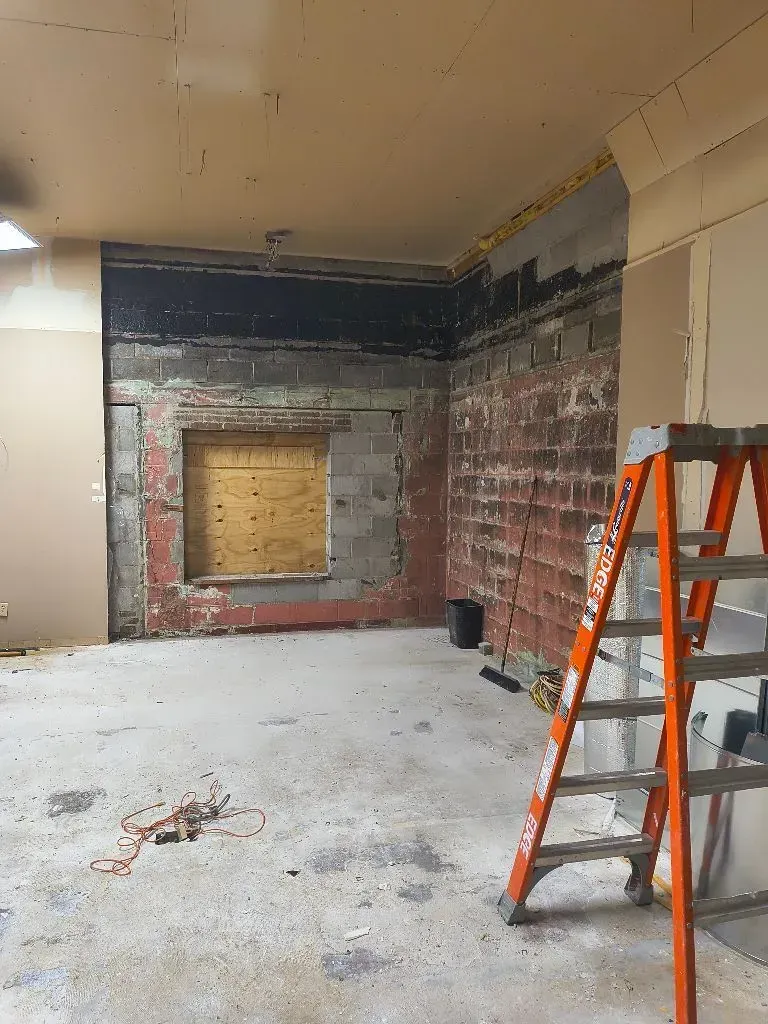 Room undergoing renovation: exposed brick wall with boarded-up window, orange ladder, concrete floor.