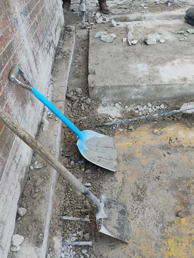 Two shovels on the ground next to a brick wall. One has a blue handle, the other is wooden. Construction site.