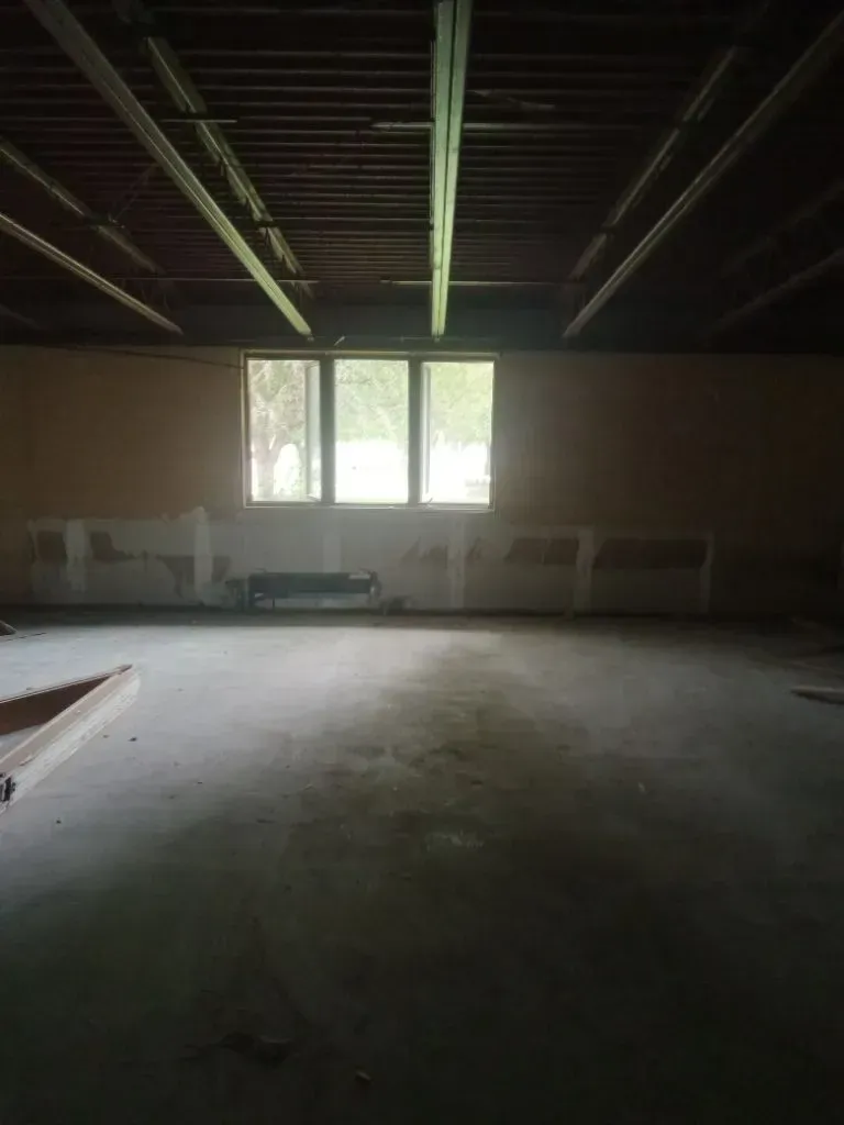 Empty room with concrete floor, three-pane window, exposed ceiling beams, and light-colored walls.