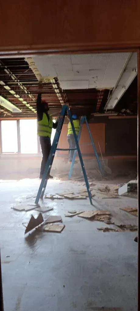 Two workers in safety vests on ladders, removing ceiling tiles in a room with debris on the floor.