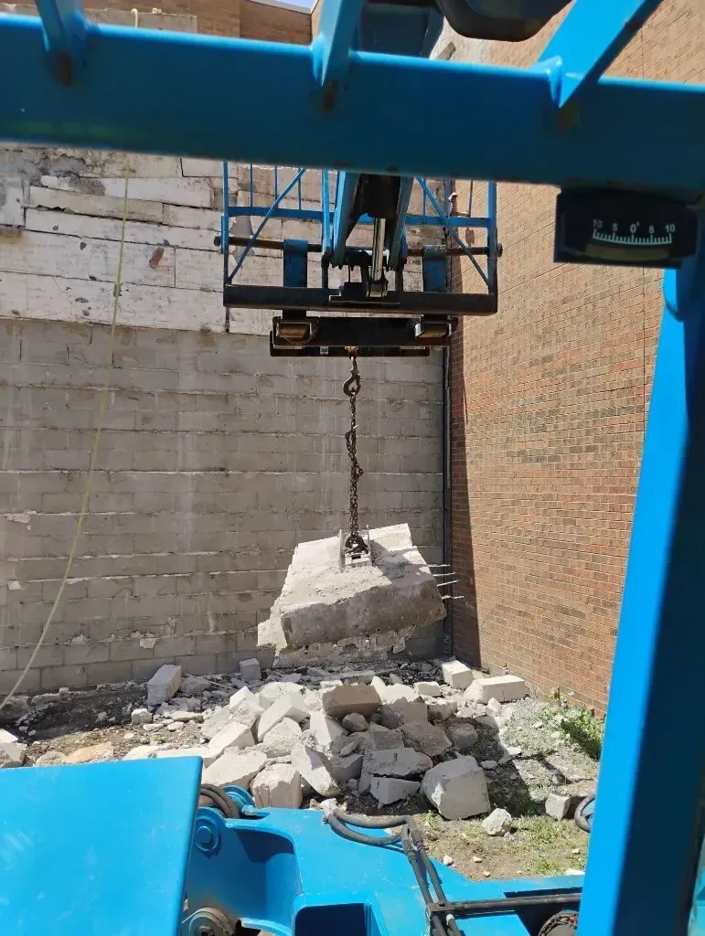 A crane lifts a concrete block from a pile of rubble in front of a brick and concrete wall.
