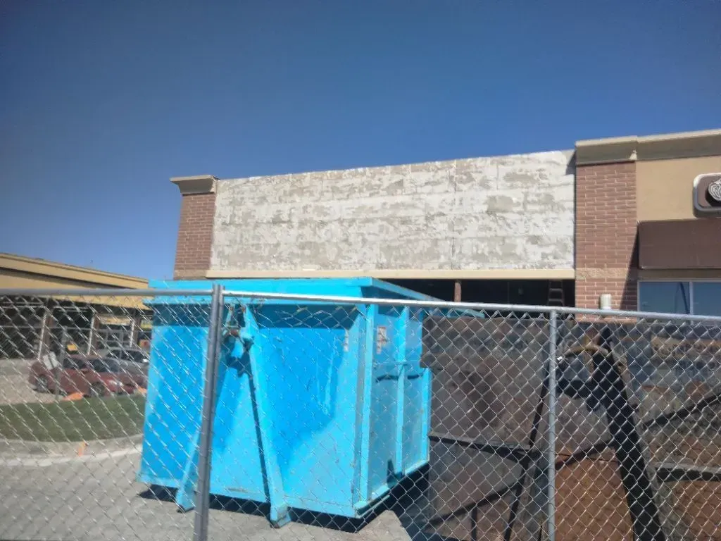 Blue dumpster in front of a boarded-up building with brick trim, behind a chain-link fence on a sunny day.